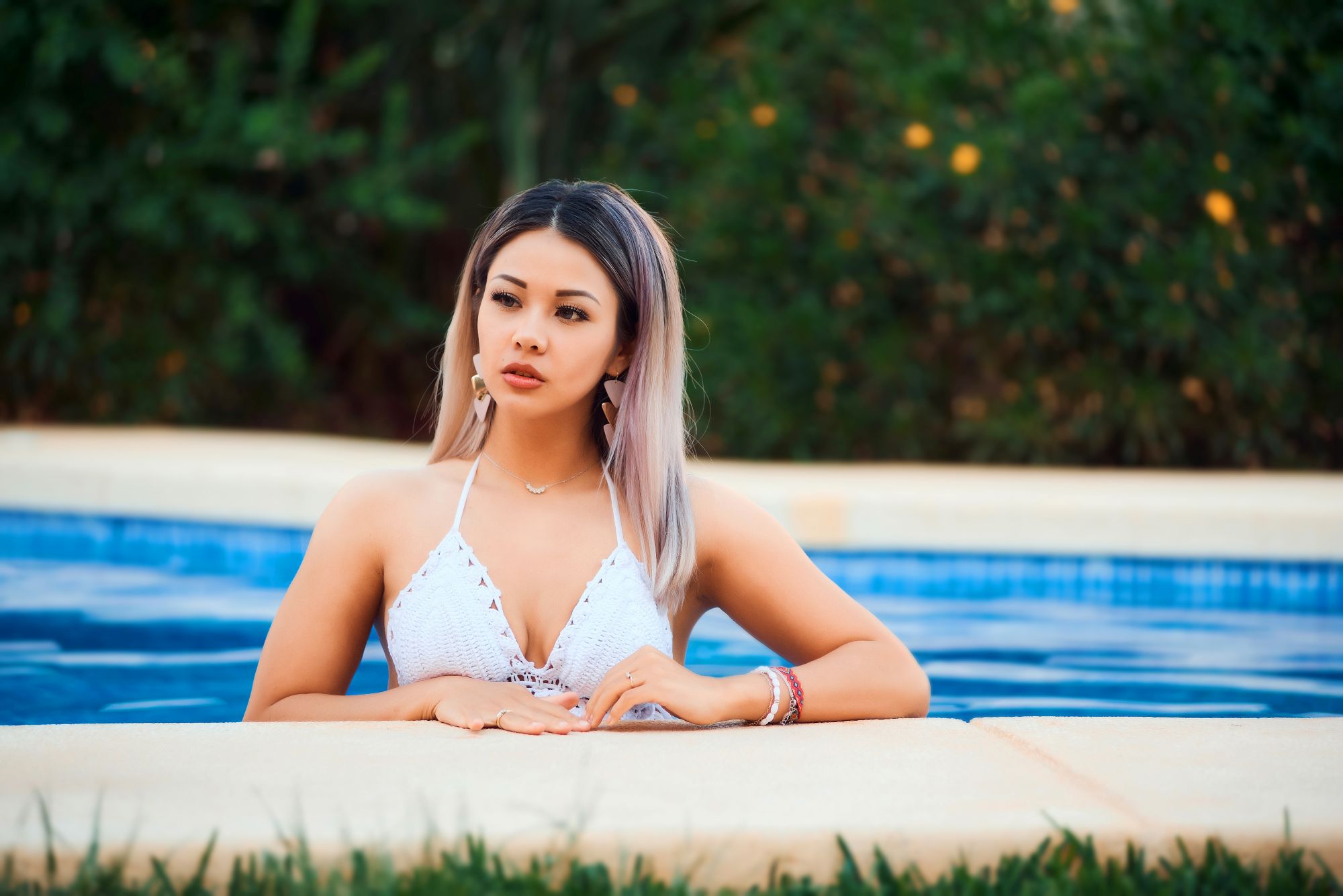 Angelic woman with blonde-highlighted hair in a white crochet bikini top leaning on the edge of a blue swimming pool